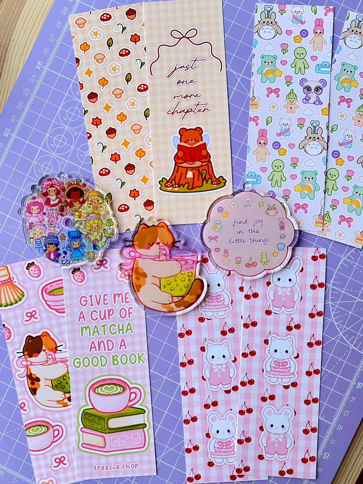 A flat lay photograph of a collection of bookmarks and keychain charms, featuring bears, bunnies, cats, mushrooms, flowers, clouds, fruit, and trinkets in various pastel tones.